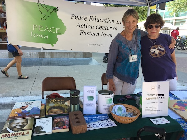 Georgiane and Nancy standing outdoors behind a table containing books, bumper stickers, and buttons. A banner with the PEACE Iowa logo hangs behind them.